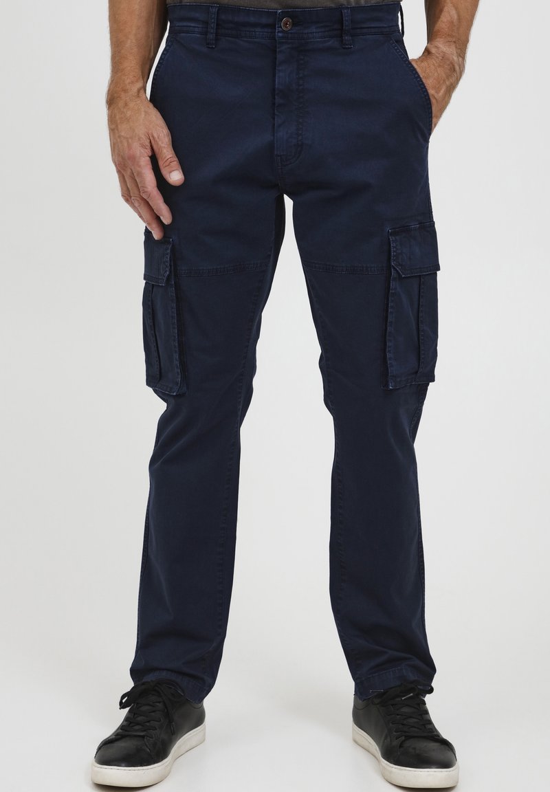 Homme portant un pantalon cargo bleu marine avec poches latérales et des baskets noires, debout les mains dans les poches devant un fond uni.