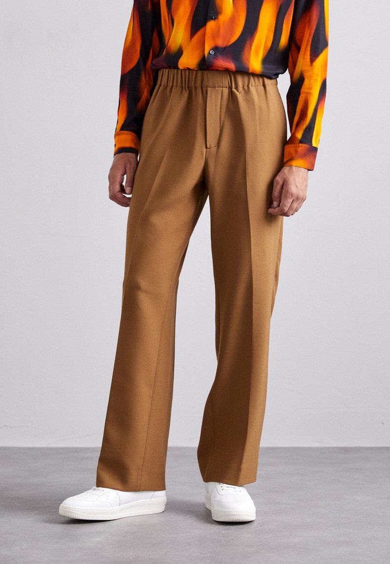 Hnědé kalhoty s volným střihem, které mají elastický pas a hladký přední díl. Doplněné o bílé tenisky na šedém pozadí.