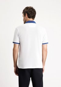 Camisa polo blanca con cuello azul y acentos en las mangas, hecha de un tejido texturizado. Presenta una pequeña etiqueta rectangular en la parte posterior.
