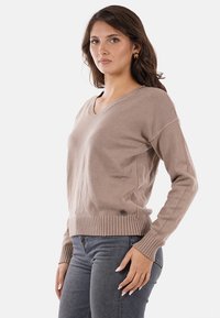 Maglione beige a V in tessuto morbido, con polsini e orlo a costine, e un discreto logo in basso. Abbinato a jeans grigi.