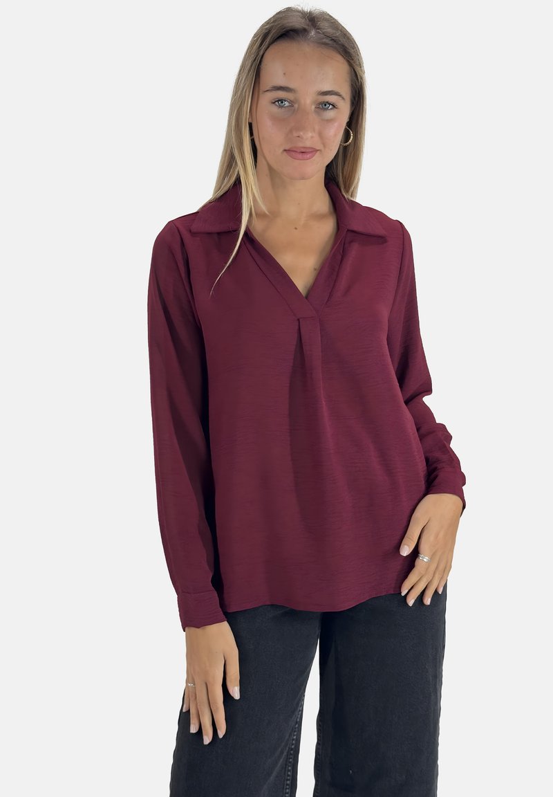 Bordeaux blouse met lange mouwen, een kraag, subtiele textuur en een plooidetail aan de voorkant. Gecombineerd met zwarte broeken.