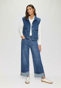 Denim vest in donkerblauw, met twee voorzakken en een knoopsluiting, gecombineerd met wijd-uitlopende donkerblauwe jeans met contrasterende boorden en witte sneakers.