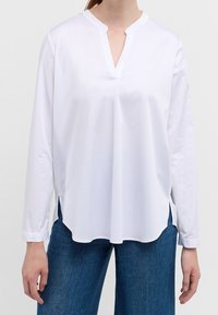 Chemise blanche à manches longues avec un col en V, des fentes latérales et une texture lisse, associée à un jean en denim bleu. Design simple et épuré.