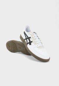 Chaussures de sport blanches en cuir et matériaux synthétiques. Elles comportent des rayures noires, un bout perforé et une semelle en caoutchouc avec une adhérence texturée.