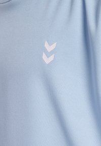 Jasnoniebieska tkanina z dwoma małymi białymi chevronami skierowanymi w dół, umieszczonymi na środku powierzchni, pokazująca teksturę tekstyliów i lekkie fałdy.