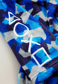 Fleecefilt i blå och grå kamomönster, med vita broderade trianglar, cirklar och fyrkantiga former som detaljer. Mjuk textur.