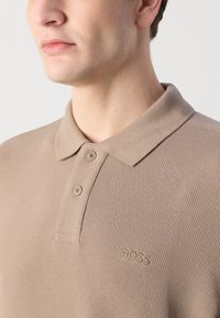Beige ribbestrikket polo skjorte med krage, to knapper og subtilt brodert merke på brystet.