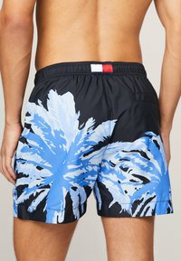 Uomo che indossa pantaloncini da bagno neri con una grande stampa di foglie di palma blu e bianche, visto di spalle, senza camicia visibile.