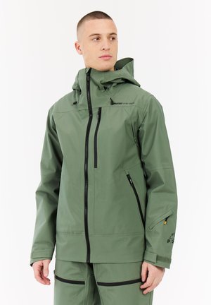 Man in een groene waterdichte jas en bijpassende broek met zwarte ritsaccenten, kijkend opzij tegen een witte achtergrond.