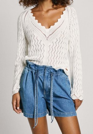 Woman wearing a white knitted long-sleeve top with scalloped neckline and high-waisted blue denim shorts with a tie belt.