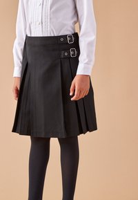 Zwarte geplooide rok met twee zilveren gespen op de tailleband, gecombineerd met donkere leggings en een witte button-up shirt.