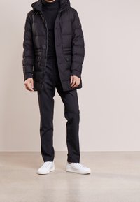 Man som bär svart vadderad jacka, svart polotröja, svarta byxor och vita sneakers, står mot en enfärgad bakgrund.