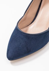 Escarpin en daim bleu marine à bout pointu avec une doublure intérieure lisse et un talon fin. Couture discrète le long du bord pour un détail supplémentaire.