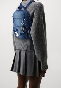 Dżinsowy plecak w niebieskim kolorze, z motywem Myszki Miki, ozdobiony srebrnymi ćwiekami, przednimi kieszeniami na zamek oraz detalem w postaci breloczka.