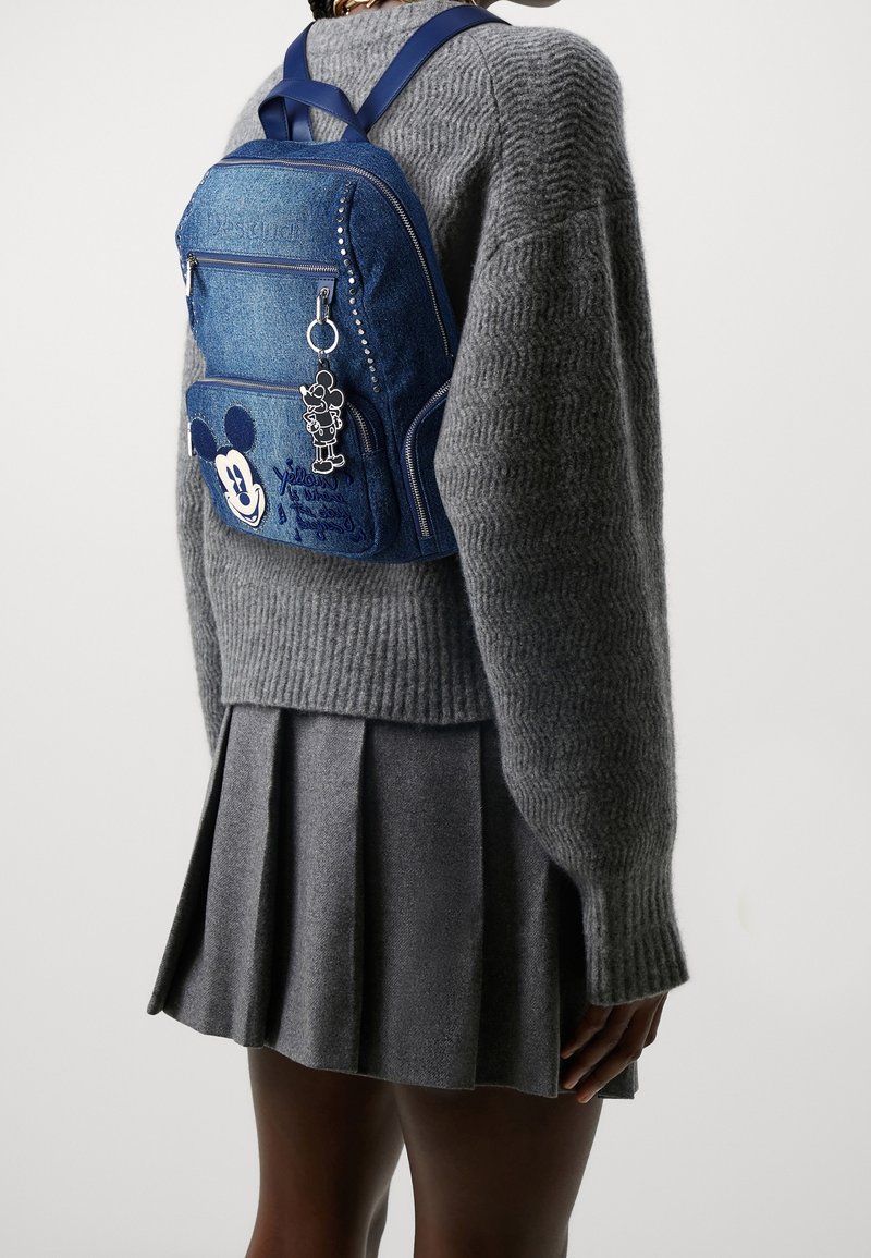Dżinsowy plecak w niebieskim kolorze, z motywem Myszki Miki, ozdobiony srebrnymi ćwiekami, przednimi kieszeniami na zamek oraz detalem w postaci breloczka.