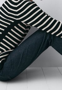 Zwart-wit gestreepte gebreide trui met een ribbelstructuur, in combinatie met een donkere spijkerbroek. De focus ligt op het bovenste kledingstuk en de positie van de arm.