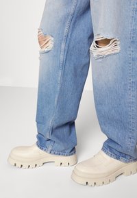 Světle modré roztrhané džíny kombinované s masivními krémovými platformovými botami na jednoduchém bílém pozadí.