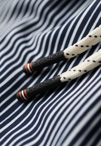 Tejido a rayas en azul marino y blanco con superficie texturizada; cuenta con cordones blancos con acentos en negro y naranja y la marca "SUPERDRY" en los extremos.