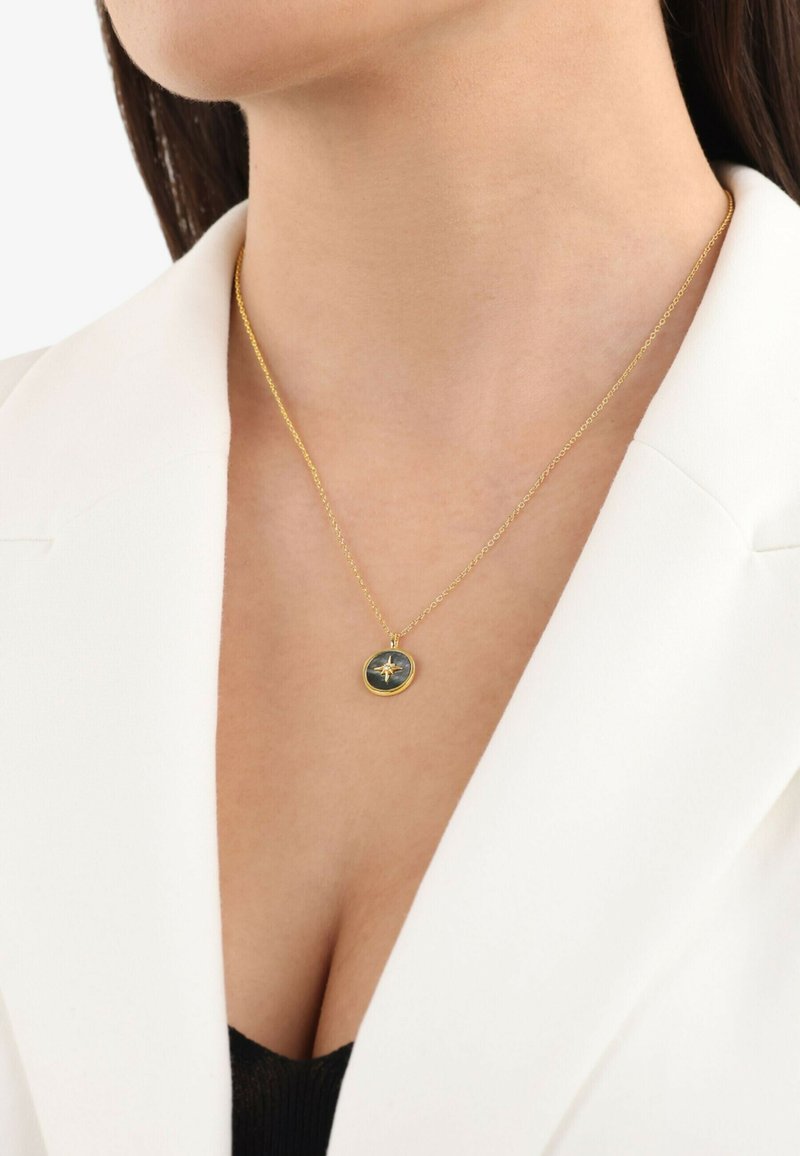 Collana d'oro con un pendente circolare scuro raffigurante un design di una bussola, indossata su una maglietta nera e un blazer bianco. Catena semplice.