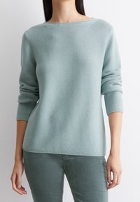 Světle modrý, texturovaný pletený svetr s lodní výstřihem a tříčtvrtečními rukávy, v kombinaci s odpovídajícími zelenými kalhotami.