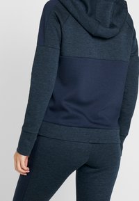 Sudadera deportiva azul marino con un acabado texturizado. Presenta una capucha, mangas largas y un panel de color contrastante en la parte superior de la espalda y en las mangas.