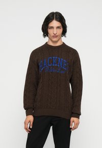 Bruine gebreide trui met een kabelpatroon, voorzien van blauwe tekst "HACKNEY Dalston Lane" over de borst. Ribbelboord bij de kraag en manchetten.