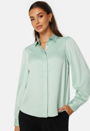 Blusa verde menta a maniche lunghe con colletto, chiusura a bottoni e spalle a sbuffo. Tessuto liscio e vestibilità comoda.