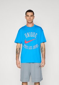 Šviesiai mėlynas per didelis T-kemšys su raudonu Nike logotipu ir tekstu "UNIQUE TOUS LES JOURS." Sukomplektuotas su pilkais šortais, turinčiais lygią tekstūrą.