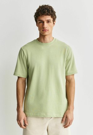 Junger Mann mit lockigem Haar, der ein hellgrünes kurzärmeliges T-Shirt und beige Hosen trägt, vor einem einfachen hellen Hintergrund stehend.