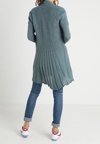 Person som bär en lång, strukturerad turkos kofta, uppvikta blå jeans och vita Nike-sneakers med röd swoosh, stående mot en enfärgad bakgrund.