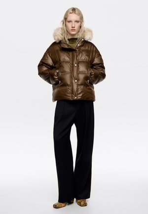 Chaqueta acolchada marrón con capucha forrada de piel, presenta un cierre de botones y dos bolsillos laterales. Combinada con pantalones de pierna ancha negros y zapatos de estampado de serpiente.