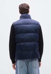 Colete acolchoado azul-marinho com gola alta, secções acolchoadas e textura suave. Usado sobre um suéter preto de malha e jeans azul claro.