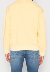 Sweat-shirt jaune clair à coupe décontractée avec des poignets côtelés, associé à un jean bleu ajusté avec des poches arrière et des détails de couture subtils.