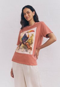Woman wearing a rust-colored T-shirt with a colorful toucan print and "GUINNESS" text, paired with light beige loose pants.