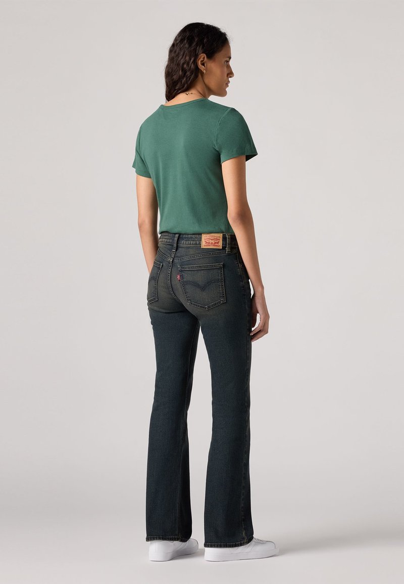 T-shirt vert à manches courtes associé à un jean évasé bleu foncé. Le jean présente une étiquette de marque rouge et jaune sur la taille. Baskets blanches.