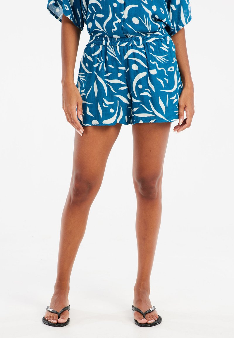 Shorts bleus avec motif abstrait blanc, taille élastique, coupe décontractée, tissu léger, associés à des tongs noires.