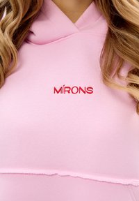 Felpa rosa con cappuccio; presenta un orlo grezzo e un testo ricamato rosso che recita "MÍRONS". Il tessuto appare morbido e elasticizzato.