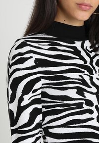 Kobieta ubrana w długą koszulę w czarno-białe paski zebry, z czarnym, prążkowanym kołnierzykiem typu mock neck i złotymi kolczykami w kształcie kół.