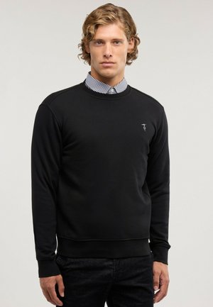 Man med ljust lockigt hår som bär en svart sweatshirt över en svartvit randig skjorta med krage, stående mot en slät ljus bakgrund.