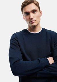 Granatowa bluza z okrągłym dekoltem, długimi rękawami, ściągaczami i gładką teksturą. Dopasowany krój o prostym wyglądzie.