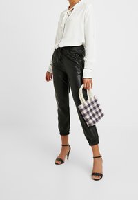 Donna che indossa pantaloni in pelle nera, camicia bianca, sandali neri con tacco alto, mentre tiene in mano una piccola borsa a tracolla in perline a scacchi.