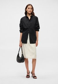 Camisa preta de botões com mangas dobradas, combinada com uma saia creme com embellishments florais. A modelo segura uma bolsa preta e usa sandálias de tiras.