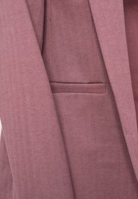 Κοντινό πλάνο τσέπης ρόζ σακακιού με σχέδιο ψαροκόκκαλο, με ορατή υφή υφάσματος και λεπτομέρειες ραφής.