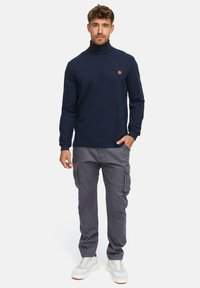 Pull torsadé marine, en tissu léger ; associé à un pantalon cargo gris avec des poches latérales, complété par des baskets blanches.