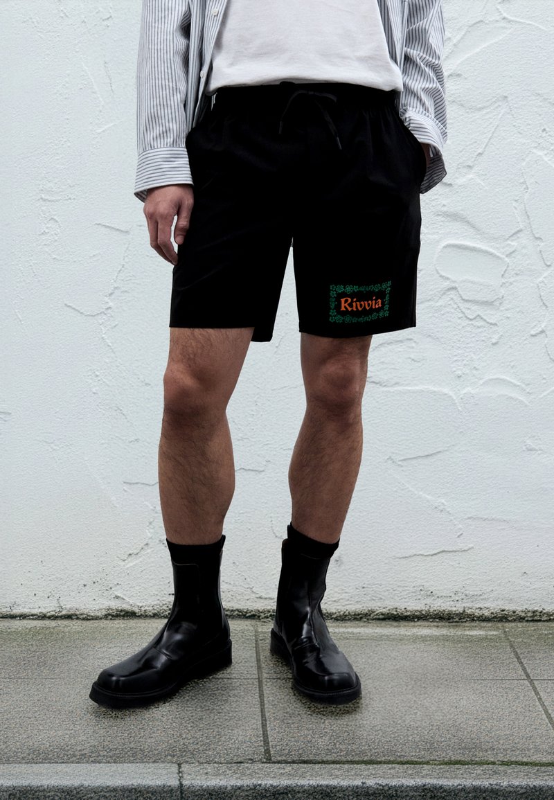 Schwarze Shorts mit elastischem Bund und Kordelzug, versehen mit einem grünen und orangefarbenen "Rivvia"-Logo, getragen mit schwarzen Stiefeletten.