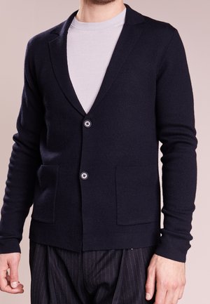 Mand iført sort knappet cardigan over hvid T-shirt med rund hals og sorte bukser med tynde striber, stående mod en ensfarvet baggrund.