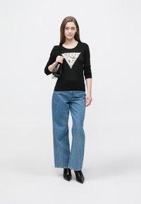 Schwarzes Langarmshirt mit einem Pailletten-Dreiecksmotiv, kombiniert mit hellblauen Jeans mit weitem Bein und schwarzen Stiefeletten mit Absatz; Modell hält eine kleine Tasche.