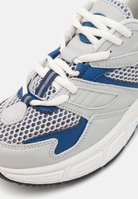 Šedá a modrá běžecká obuv s nylonovou tkaninou, koženými detaily, bílými tkaničkami a strukturovanou gumovou podrážkou. Značné designové překrytí přítomno.