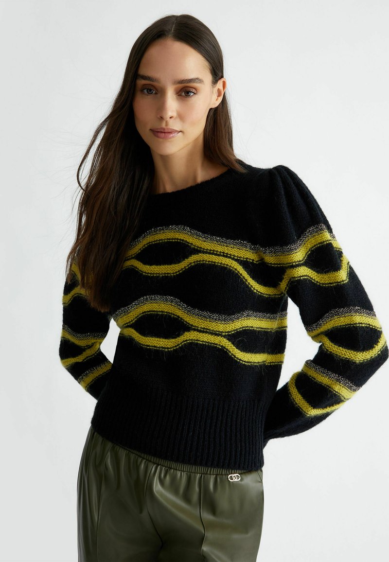 Maglione nero a maglia con strisce ondulate gialle e argento, orlo a costine e dettagli sulle maniche a sbuffo, abbinato a pantaloni verde oliva.