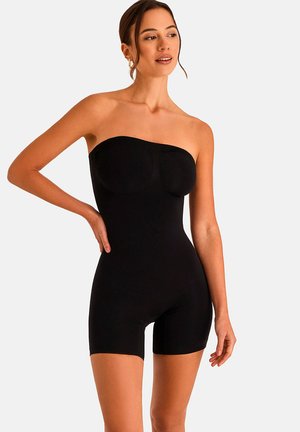 Femme portant un short gainant noir sans bretelles, debout avec une main sur la hanche, regardant légèrement sur le côté sur un fond uni.